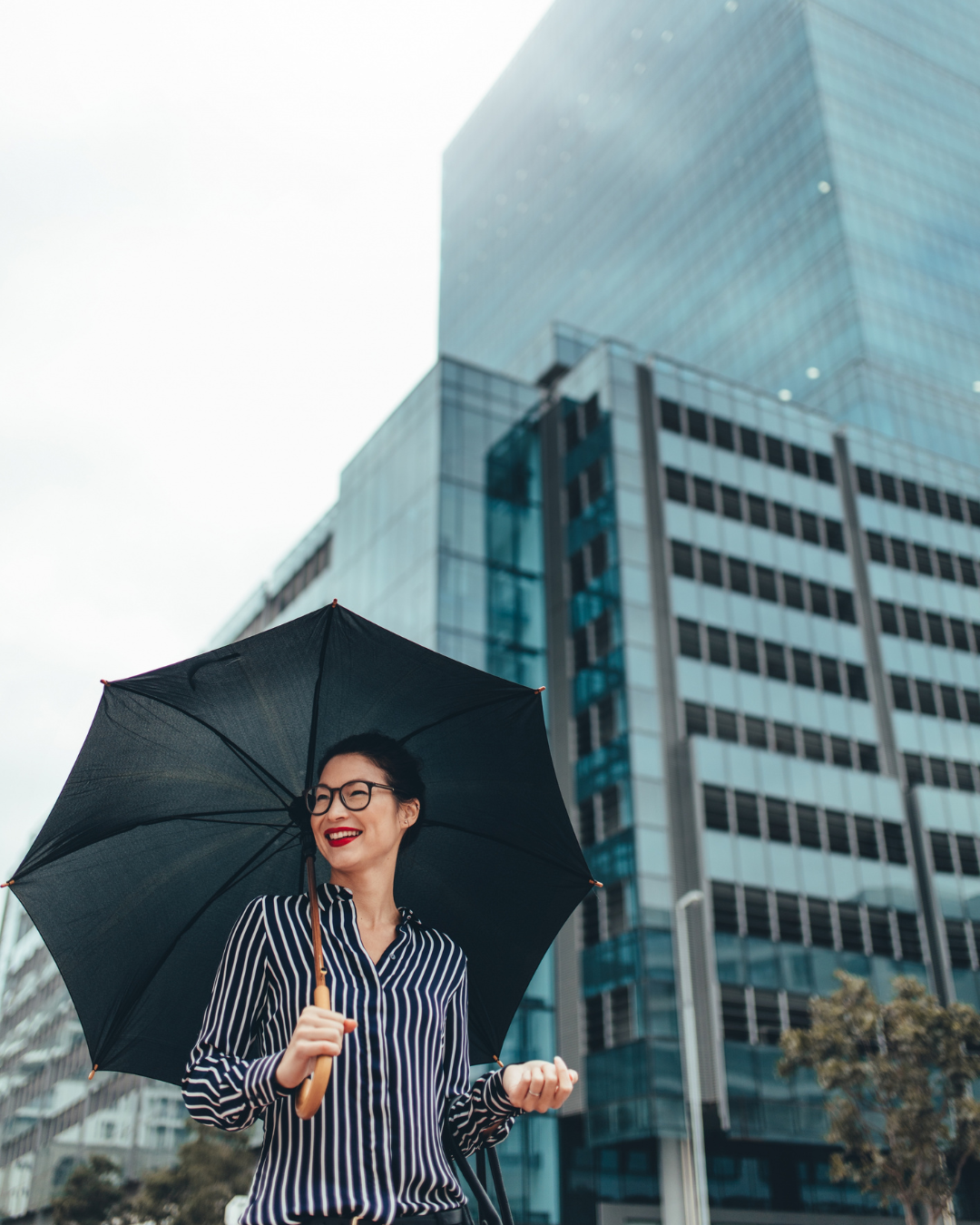 A smiling businesswoman stands in front of a large office building and protects herself from the rain with an umbrella